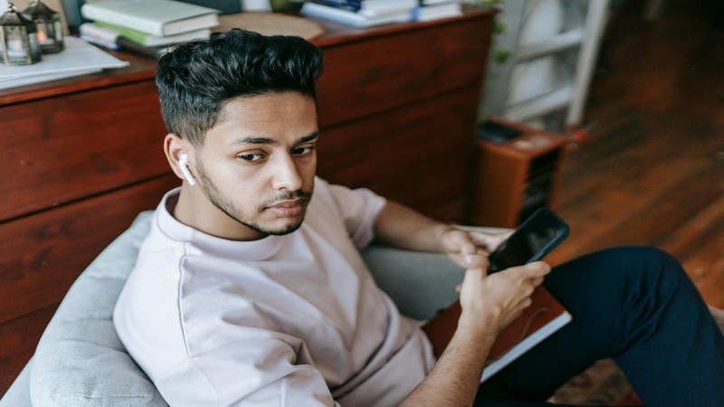Person sitting on a chair with a smartphone and wireless earbuds. Books and items are on a wooden surface behind them.